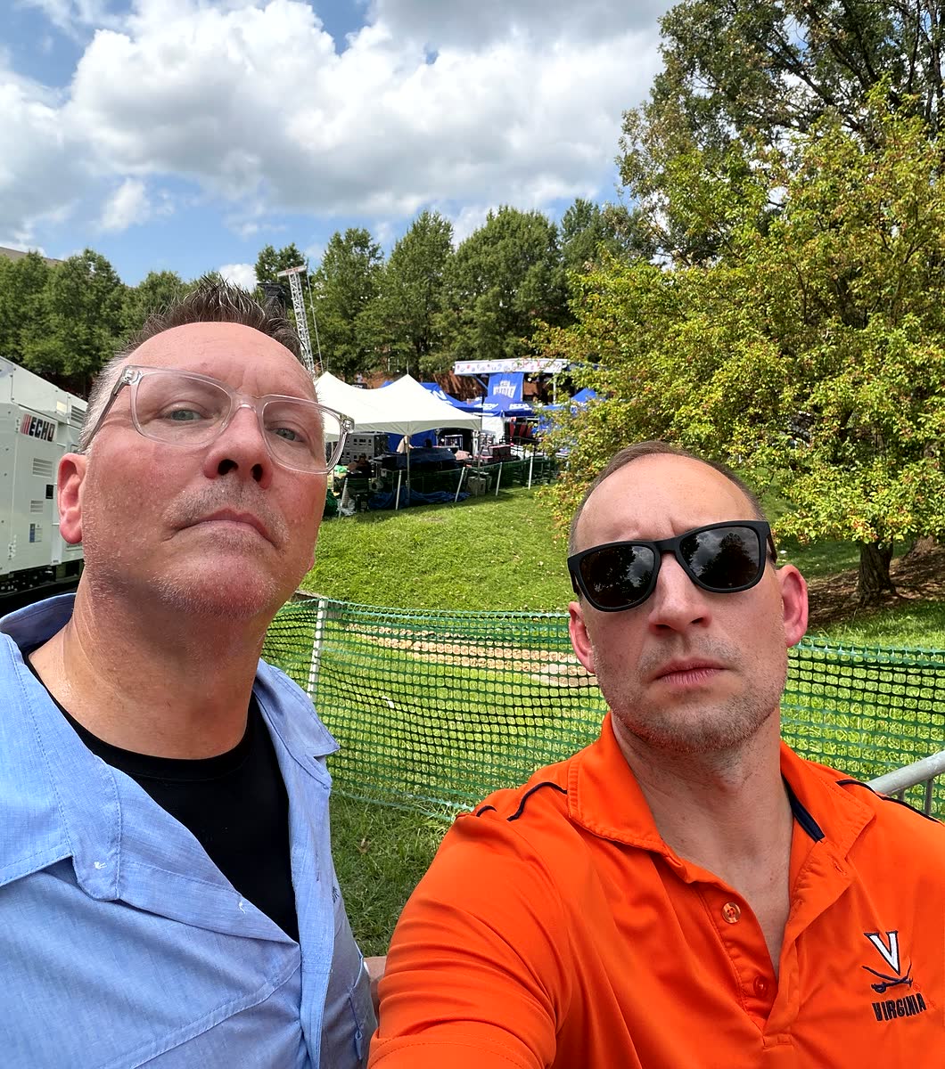 Two men taking a selfie in front of the fenced-off ACC Network broadcast compound on the lawn.