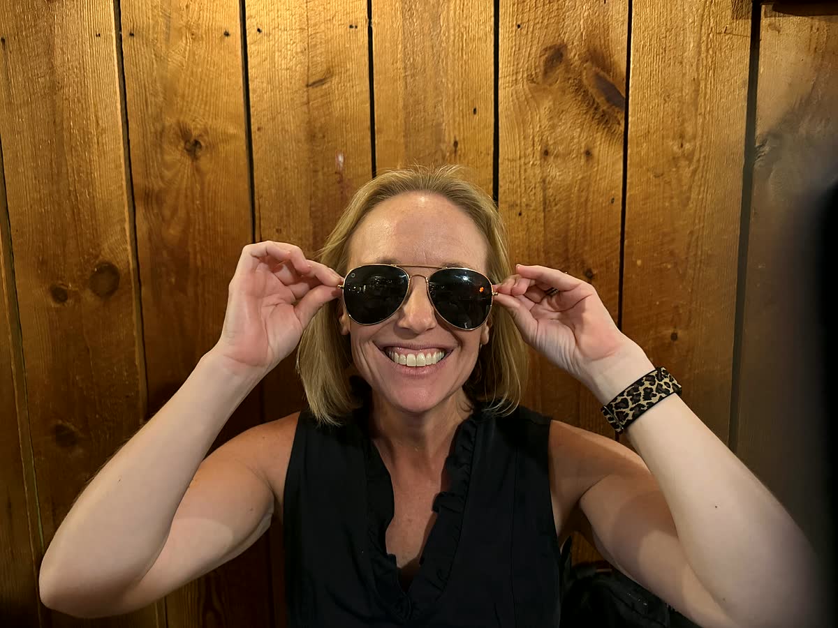 Nicole grins and holds a pair of aviator sunglasses on her face, against a wood-paneled wall.