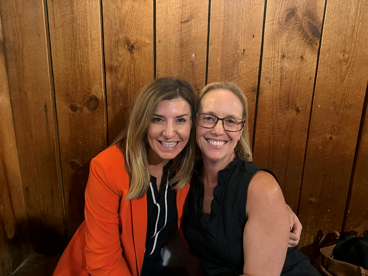 Darcie, in an orange blazer, and Nicole lean together for a photo in a wood-paneled booth.