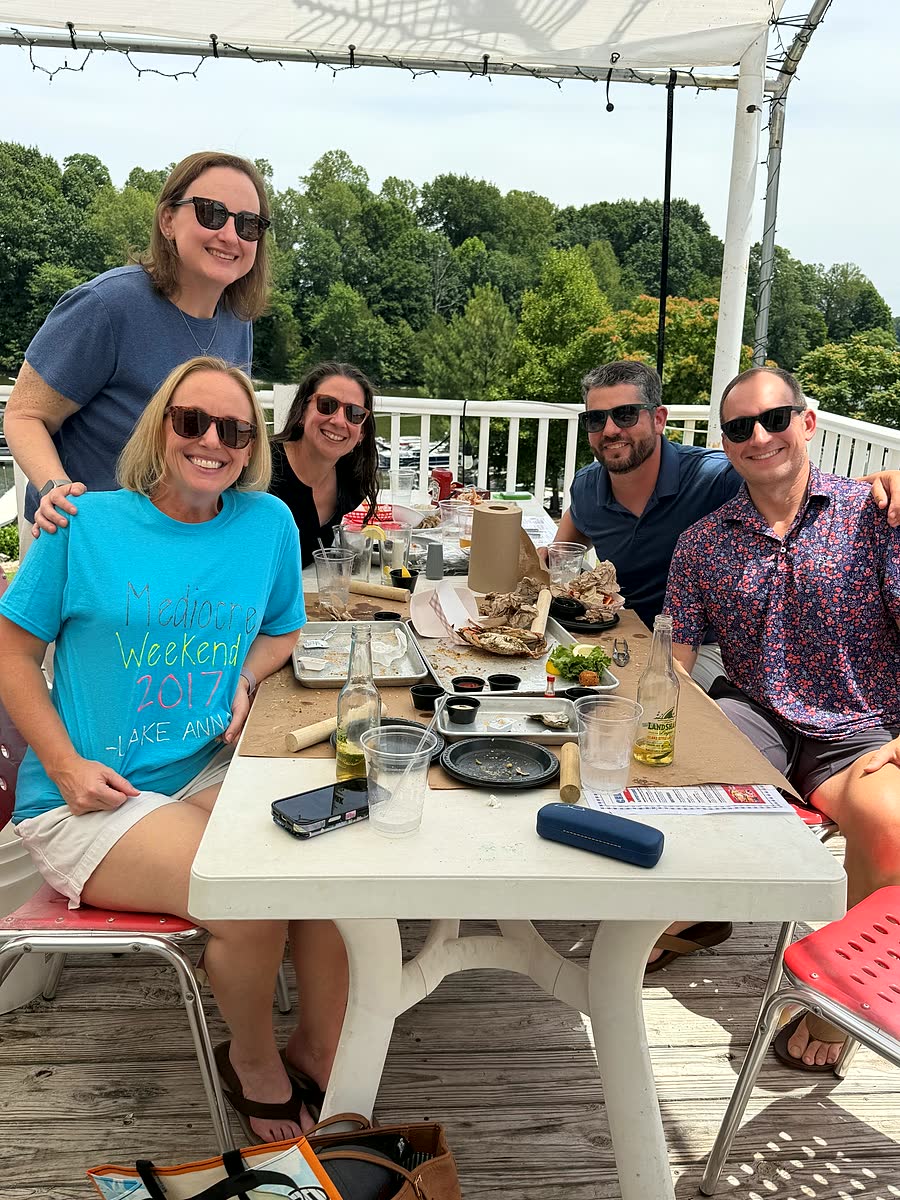 Five friends in sunglasses around a table on a sunny deck, with trees behind them.