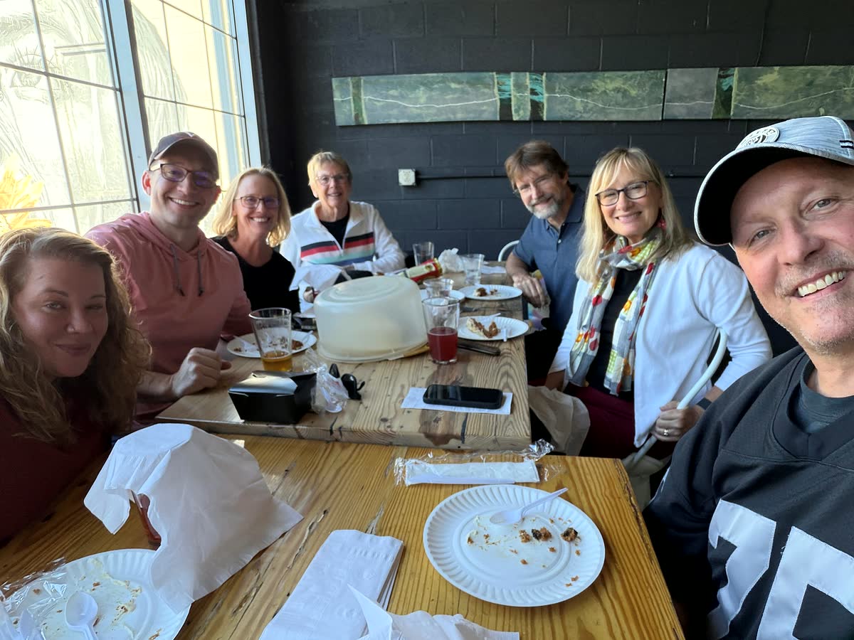 Eight people smiling around a long wooden table by a big window, over empty plates.