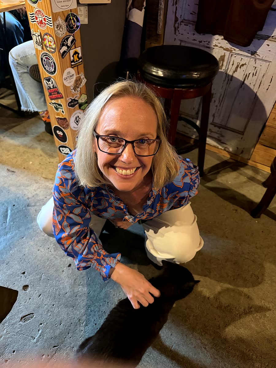 Nicole, in a blue and orange top, crouches on the concrete floor of a bar and smiles at the camera, one hand on a black cat.