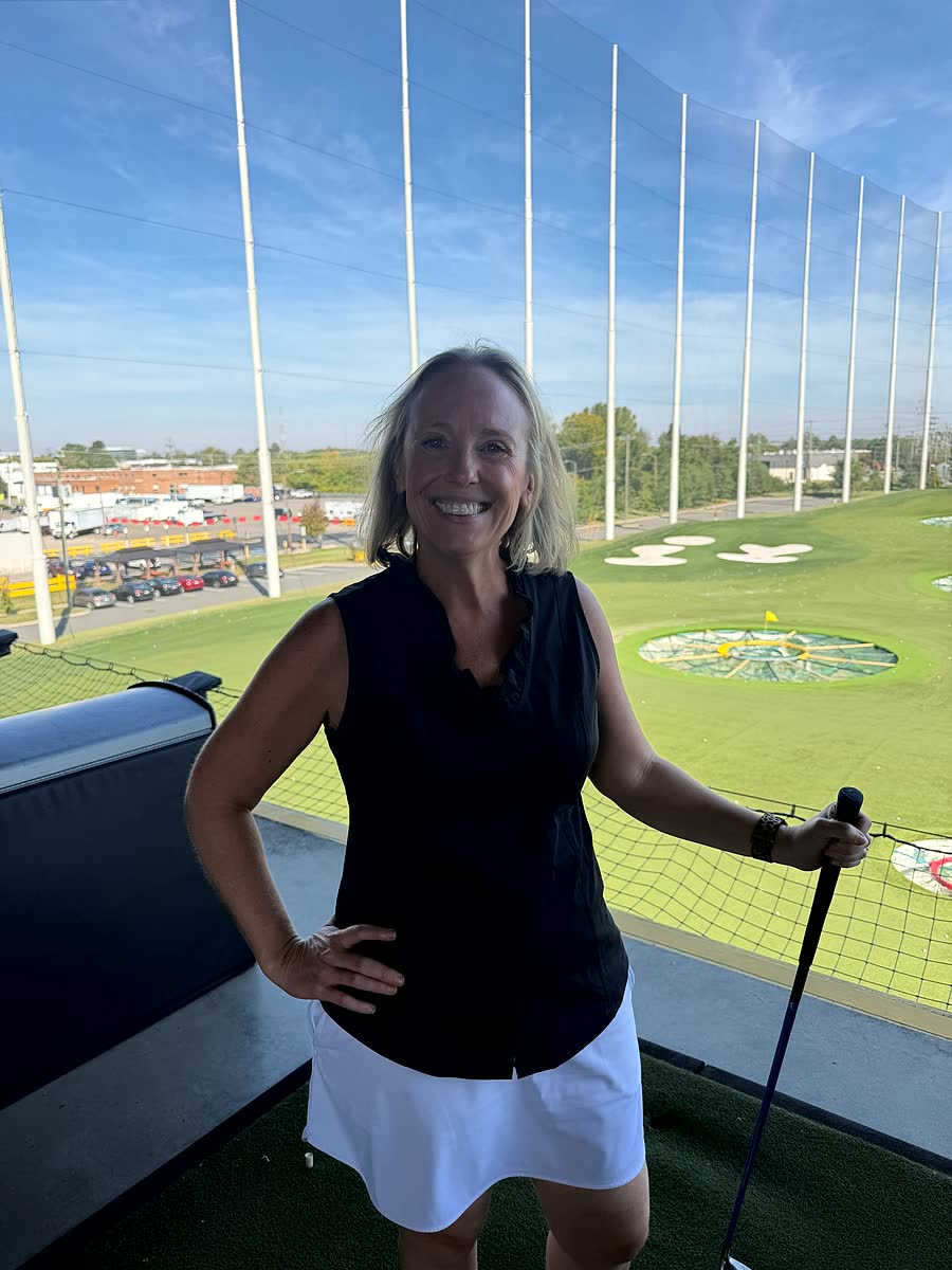 Nicole stands in a Topgolf bay holding a club, the range and its target rings behind her.