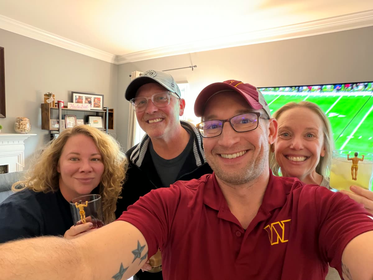 A selfie of four smiling friends in a living room. The man in front wears a burgundy Commanders polo and cap, and a football game is on the big TV behind them. Two of them hold drinks with small gold football-player figures clipped to the rims of their glasses.