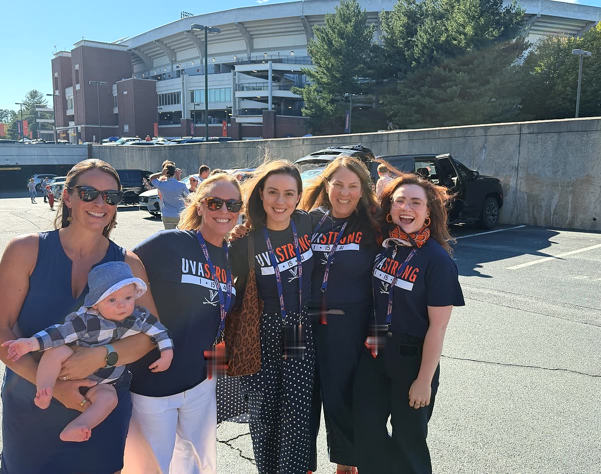 Five women standing shoulder to shoulder in a parking lot outside Scott Stadium on a bright, clear afternoon, most of them in navy UVA STRONG shirts. The woman on the left holds a baby in a checked outfit and a pale sun hat. The stadium and a line of trees rise behind them.