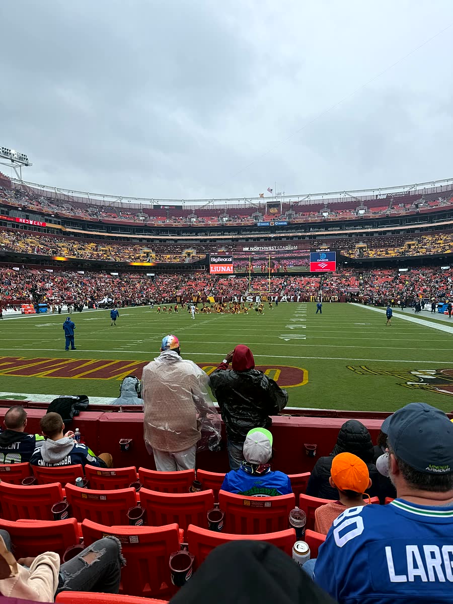 A wide view of the inside of Northwest Stadium from the stands on an overcast, rainy afternoon, with the field and far stands visible.