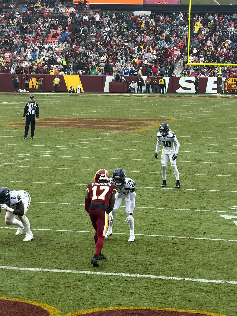 A Commanders receiver in burgundy running a route against two Seattle defenders in white, viewed from the stands.