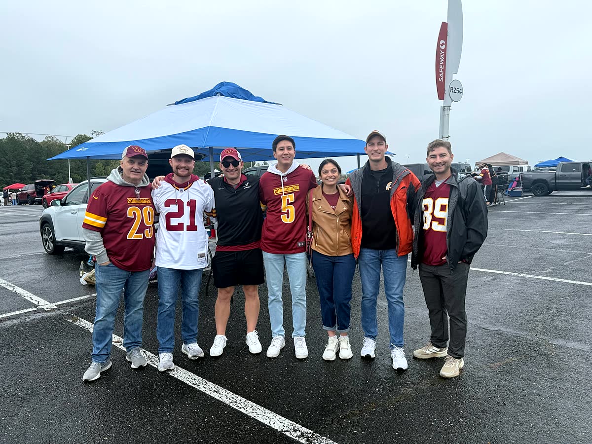 A group of seven people in Commanders jerseys standing shoulder to shoulder in a wet parking lot in front of a blue tent.