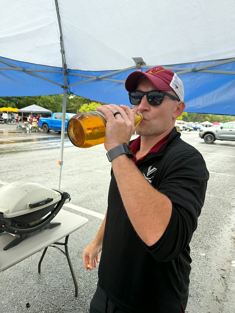 A man in a burgundy cap and sunglasses drinking from a large bottle of beer under the tent in the parking lot.