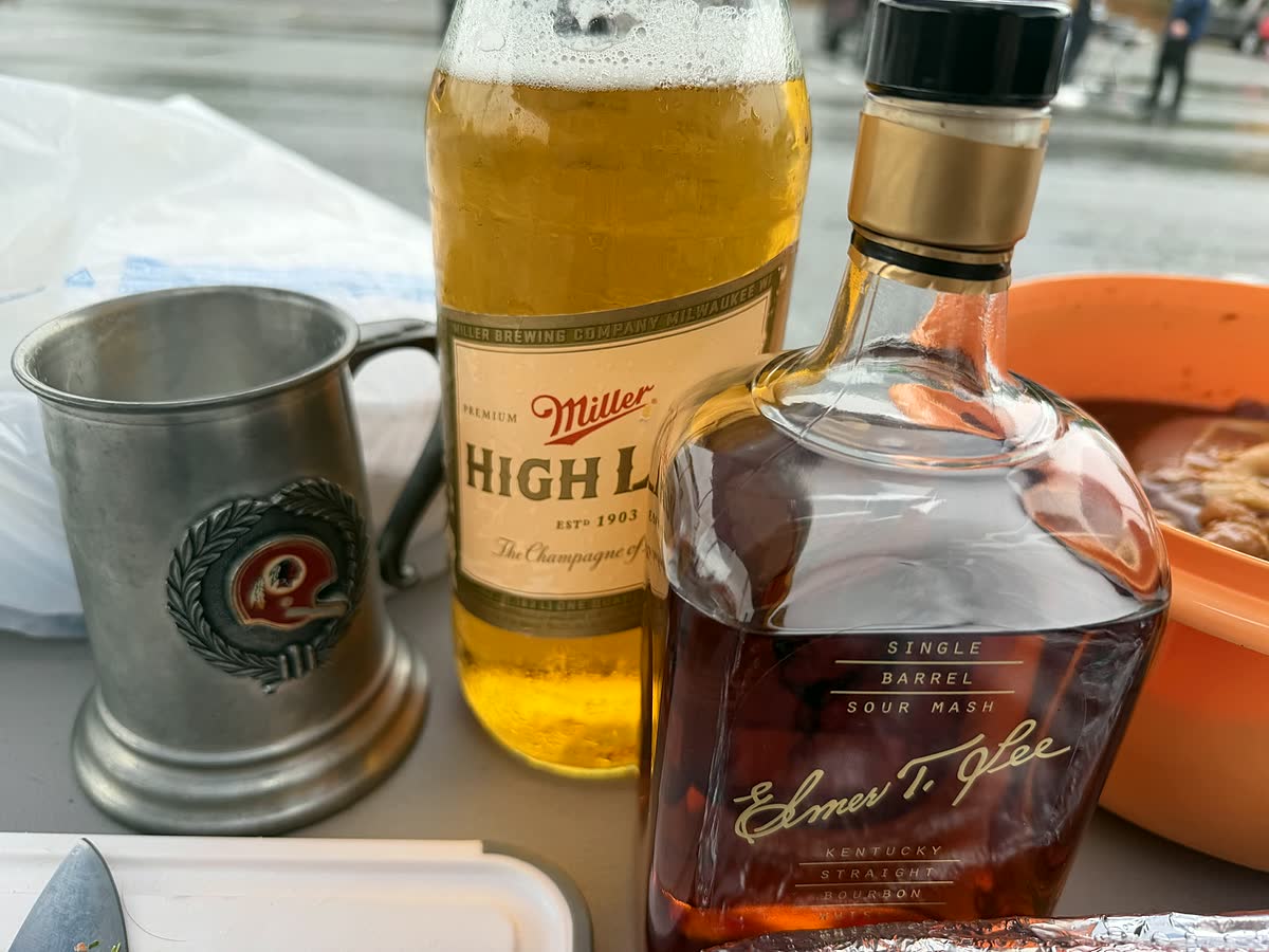 A bottle of Miller High Life and a bottle of Elmer T. Lee bourbon on a table next to an old pewter Redskins tankard.