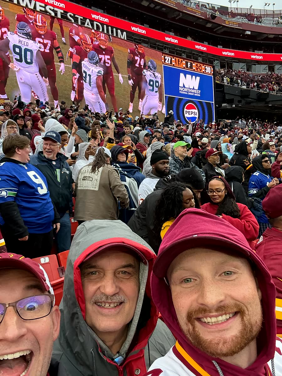 Three men in rain hoods grinning at the camera in a packed stand, with the stadium screen and crowd behind them.