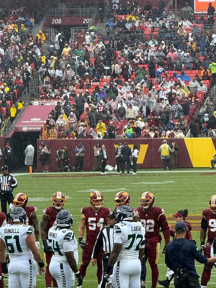 Commanders players in burgundy huddled on the field with Seattle players in white standing nearby, crowd packed behind.
