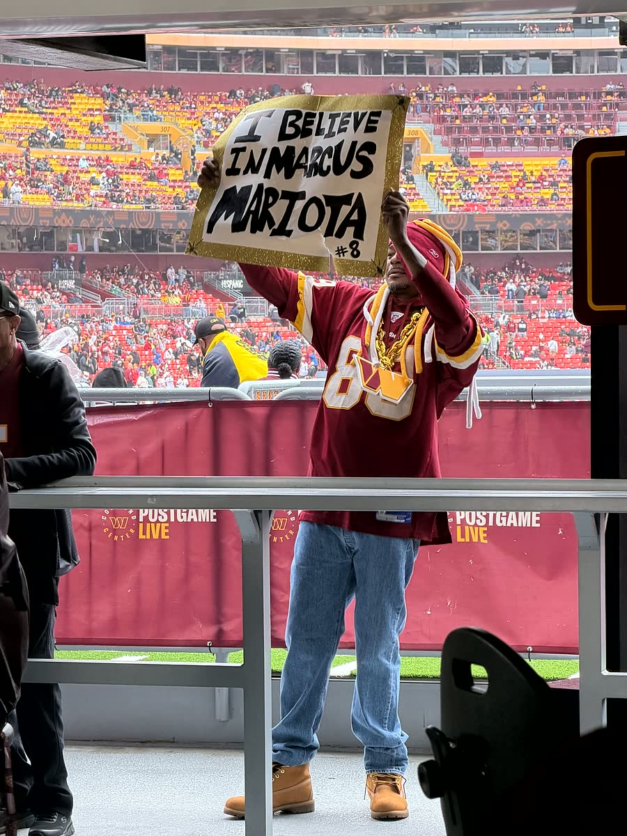 A fan in a Commanders jersey holding up a hand-lettered cardboard sign reading I BELIEVE IN MARCUS MARIOTA.