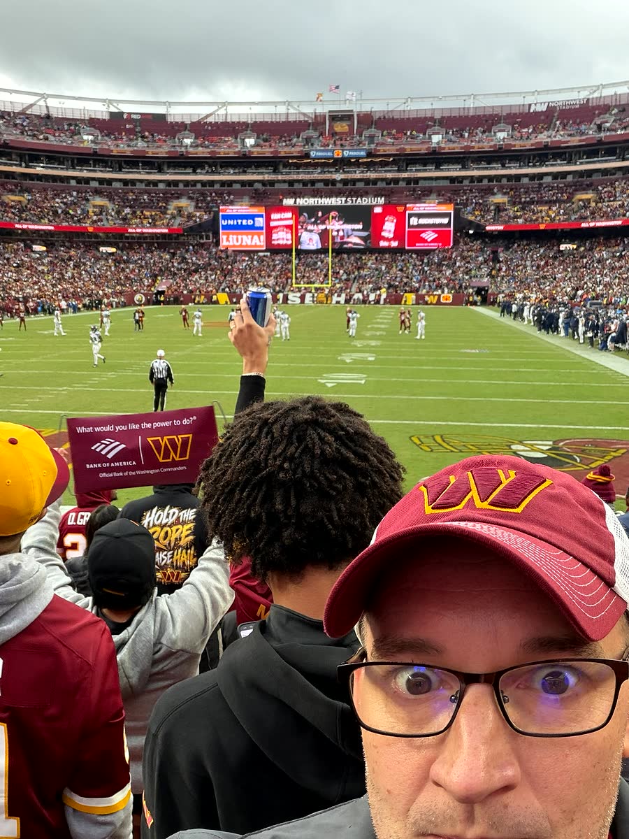 A selfie from the stands with the field and the stadium screen visible behind, crowd all around.