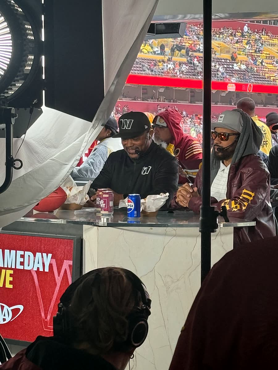 A Gameday Live broadcast position inside the stadium, with presenters at a counter and the stands visible behind.