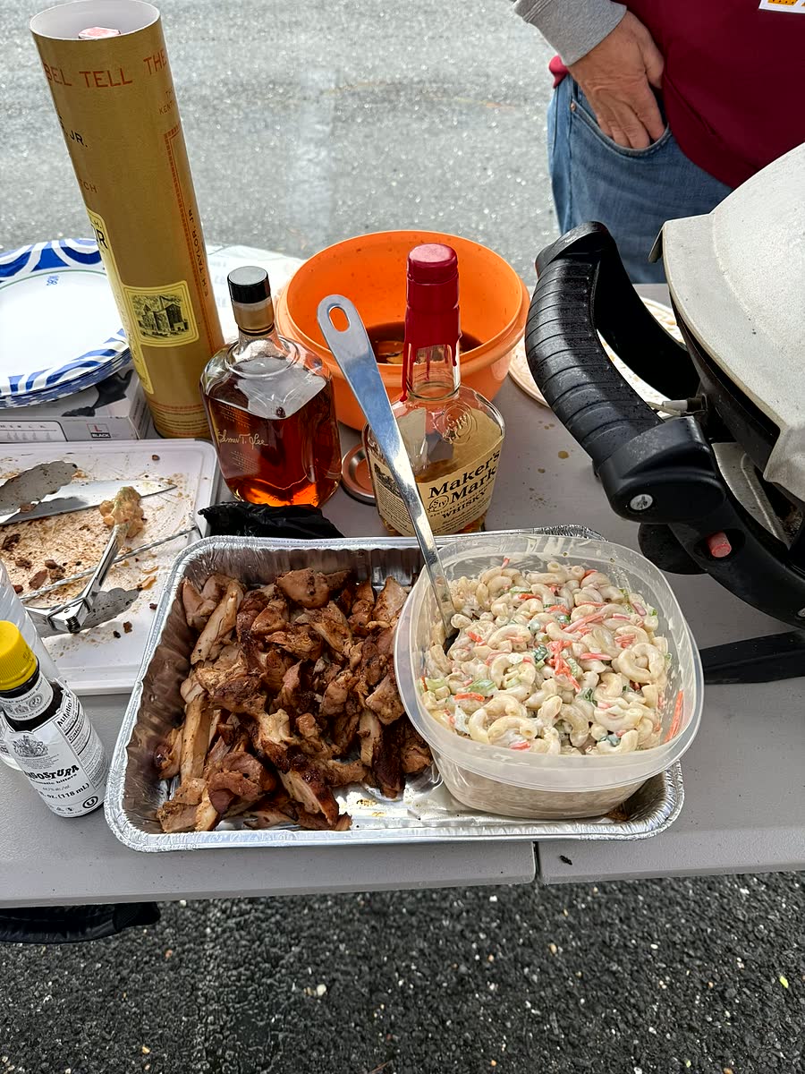 A foil tray of wings and a bowl of macaroni salad on a folding table in a parking lot, with a bottle of bourbon and an orange cooler behind them.