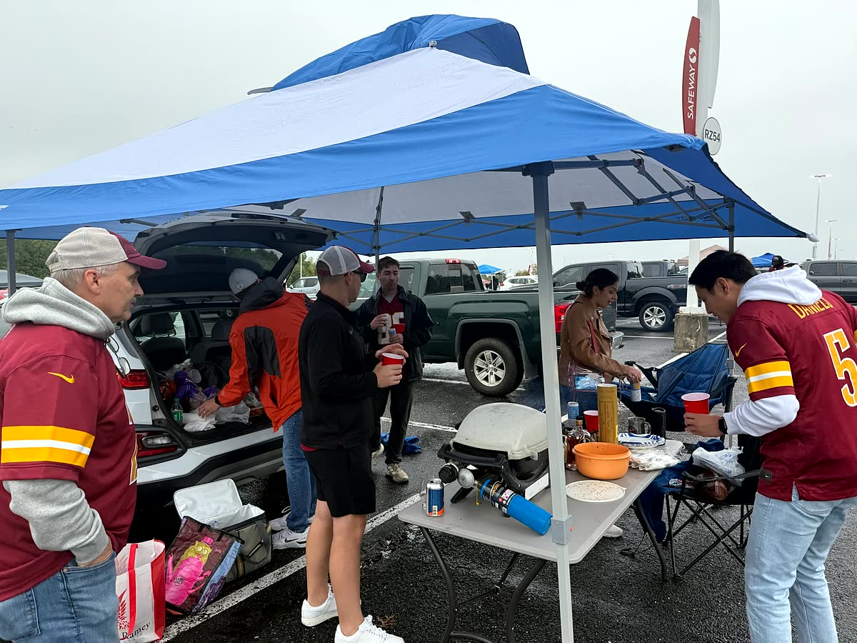 A blue pop-up tent in a wet stadium parking lot with people in Commanders jerseys setting up food on tables underneath it.