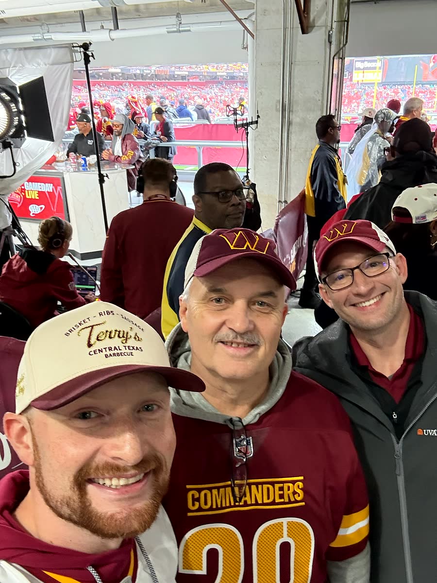 Three men leaning into the camera inside the stadium concourse, one in a Commanders jersey and cap, one older in a gray jacket, one in glasses.