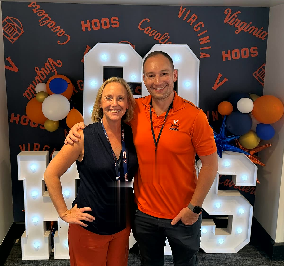 Ben and Nicole in front of a giant lit-up HOOS marquee sign with orange and blue balloons.