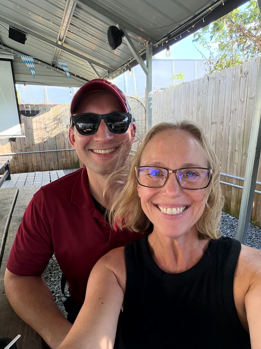 Ben and Nicole smiling in the Högwaller beer garden, with a metal roof, string lights and blue and white Oktoberfest bunting overhead.