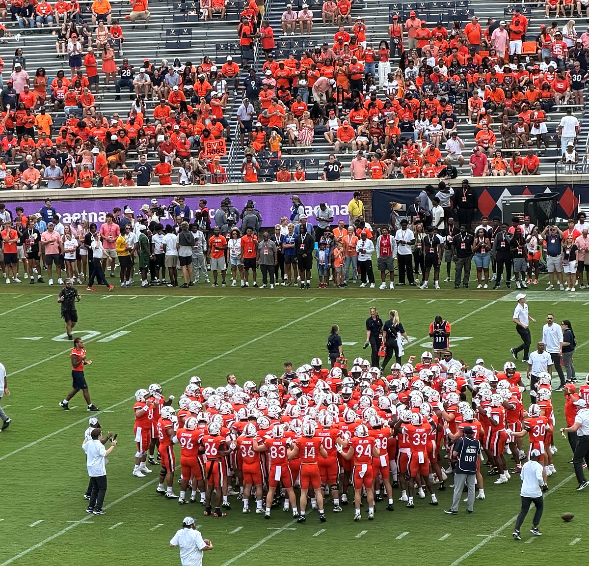 The Virginia team gathered in a tight huddle at midfield before kickoff, orange-clad crowd in the stands above.