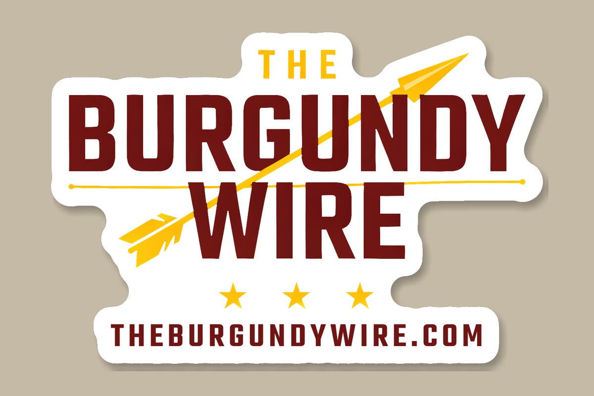 A die-cut sticker of The Burgundy Wire logo on a tan desk: burgundy letters crossed by a gold arrow, three gold stars, and THEBURGUNDYWIRE.COM along the bottom, with a thick white border cut to the shape of the logo.
