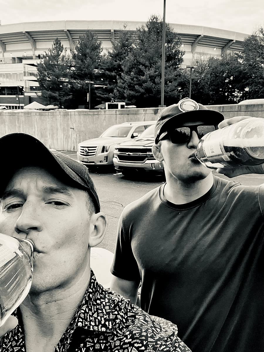 Black and white photo of two men drinking beers in a parking lot with Scott Stadium behind them.