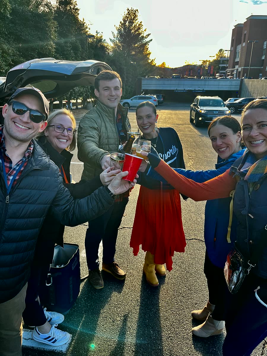 Six friends reaching in to clink beers and a red cup in a parking lot at sunset.